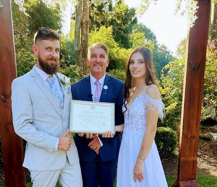 Mark and a couple holding their commemorative marriage certificate by an arbour