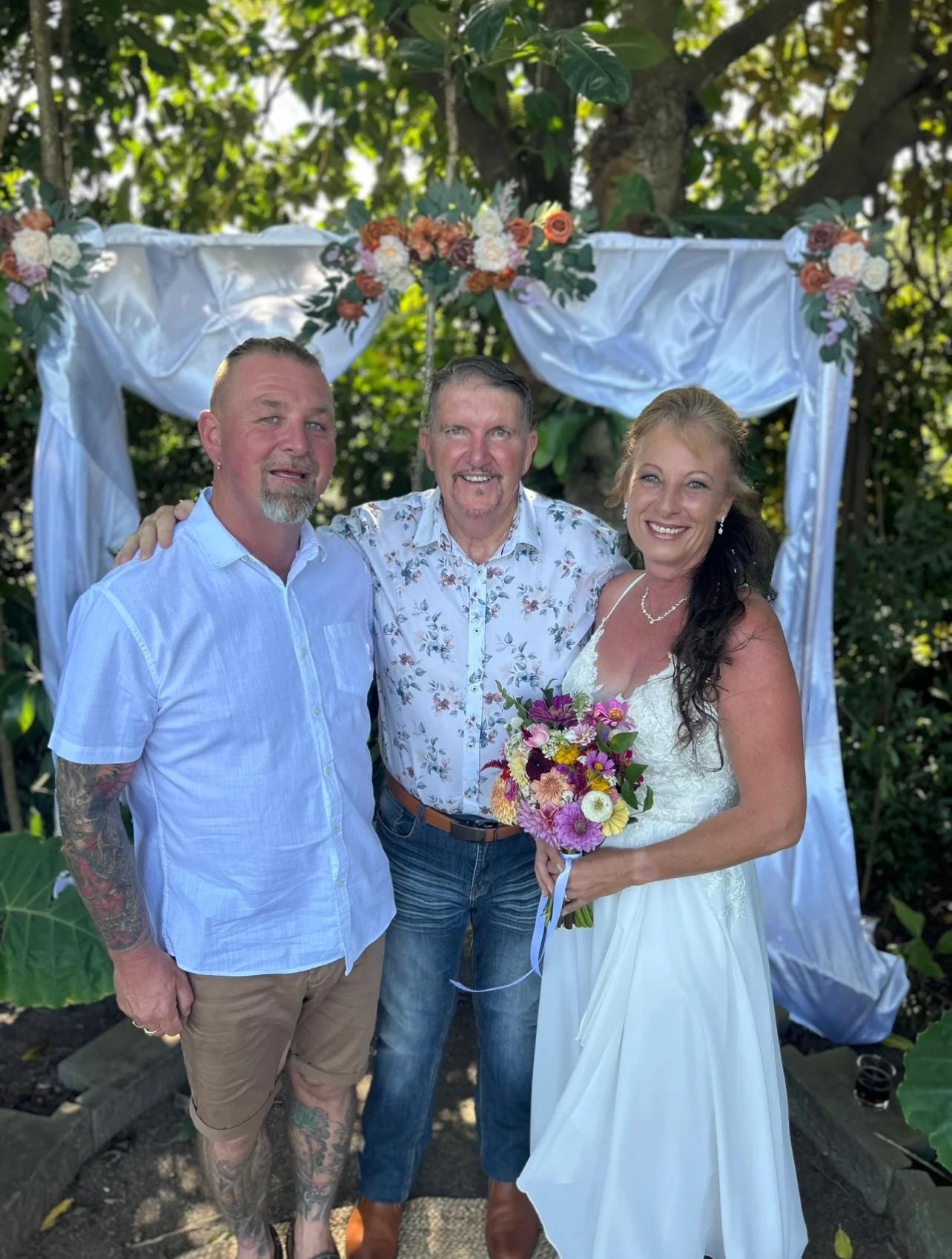 Mark standing with a couple beneath a flower-decorated arbour