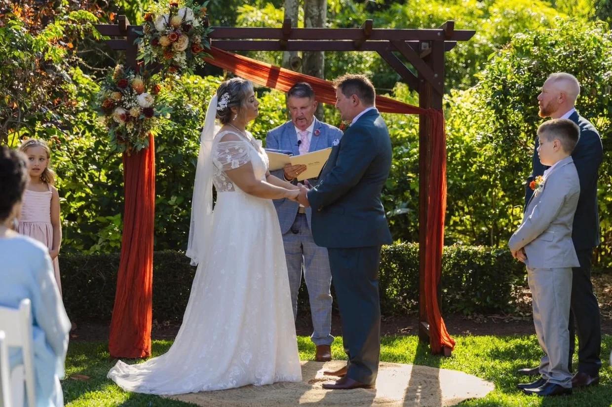 Mark officiating as a couple hold hands beneath a garden arbour
