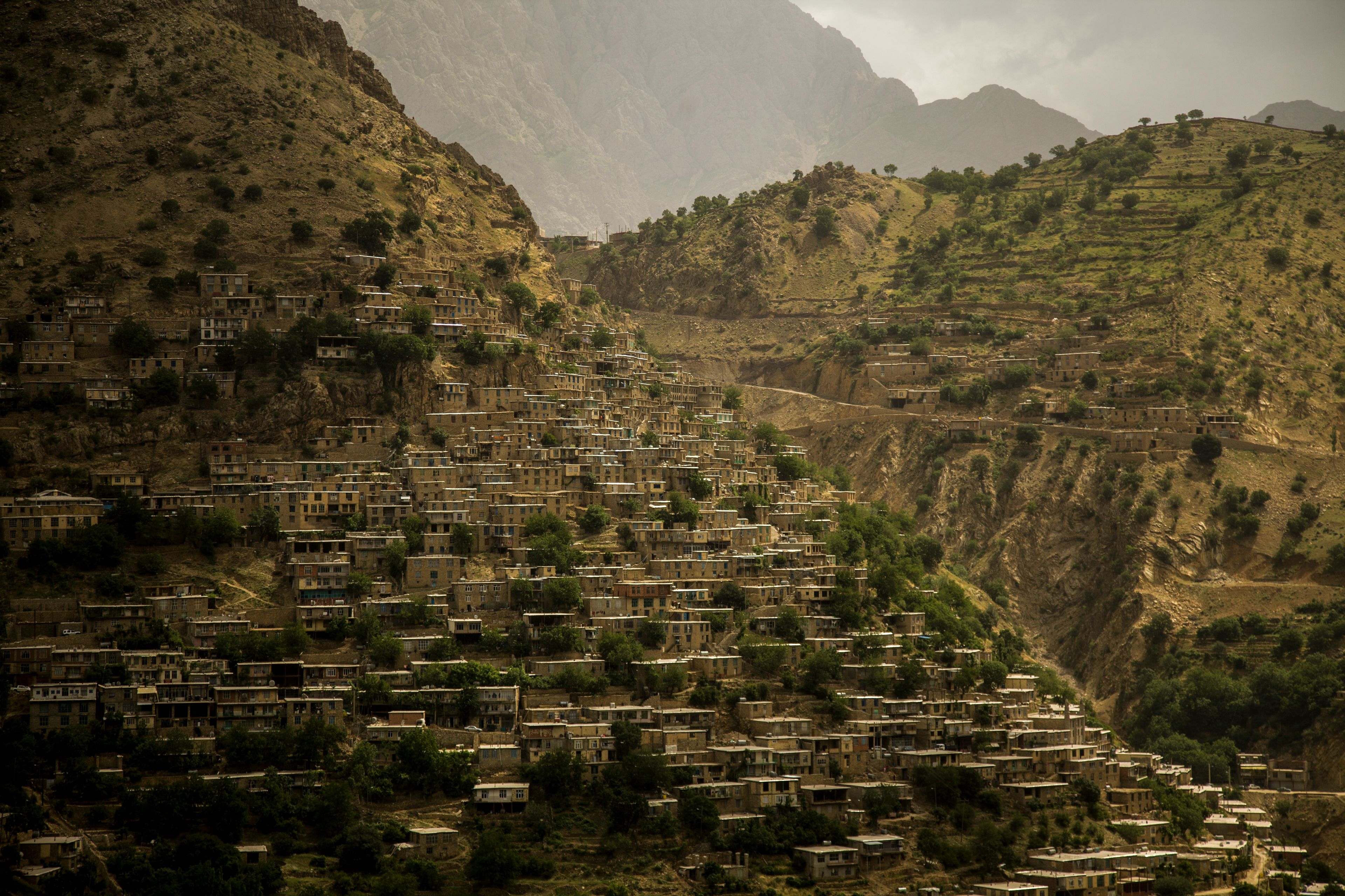 A village of flat-roofed stone houses stacked in terraces up a steep, dry mountainside, with green trees between them and terraced slopes and rocky peaks under a hazy sky behind.