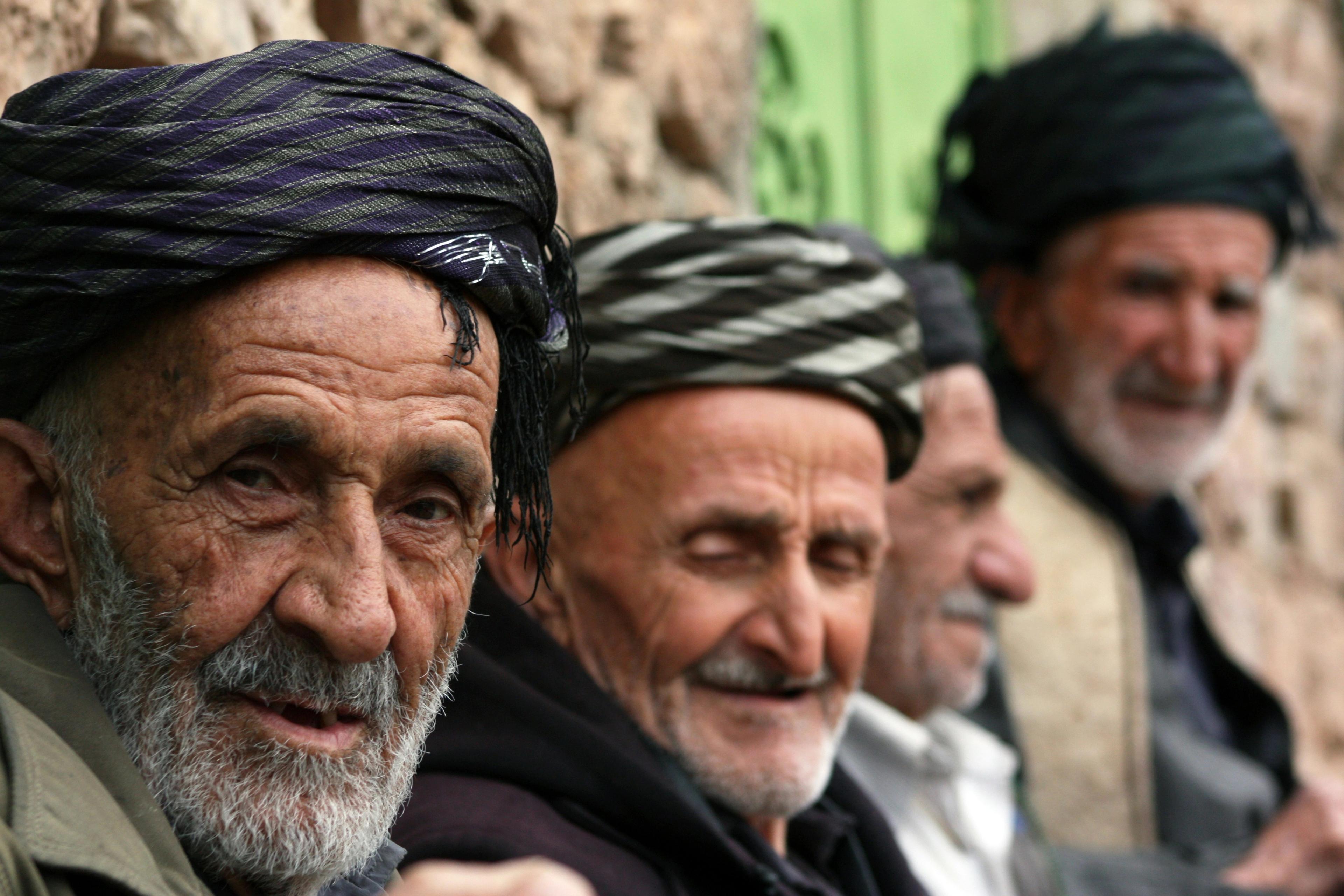 Four elderly Kurdish men in striped and dark wrapped turbans sit in a row against a stone wall; the nearest, with a white beard, looks into the camera while the others are softly out of focus behind him.