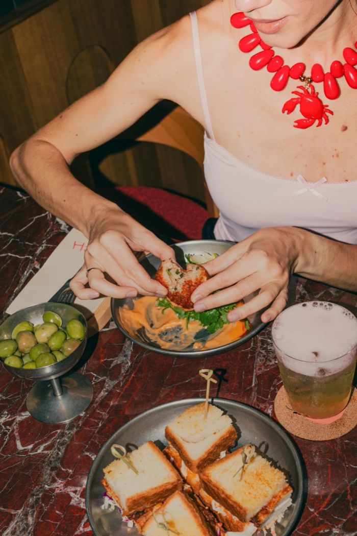 Guest eating a crispy seafood bite with a red crab necklace at Pinch Lisbon, with sandwiches and olives