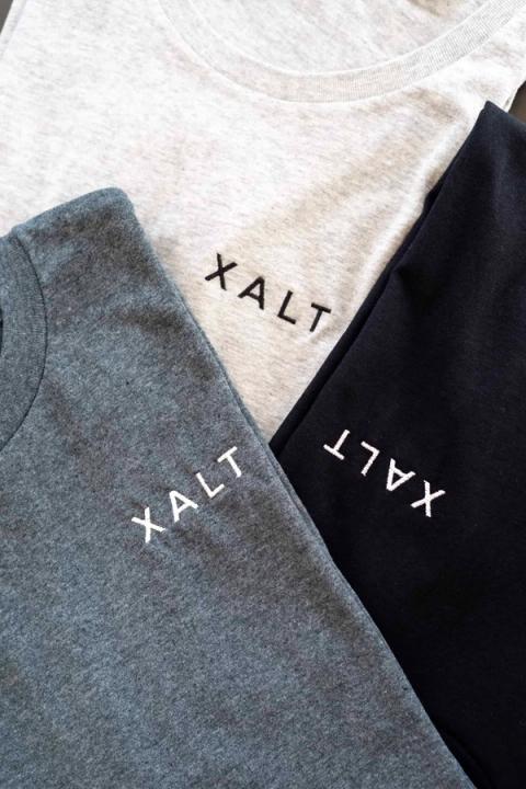 Drei übereinandergelegte T-Shirts in Hellgrau, Dunkelgrau und Schwarz mit gestickter XALT-Schrift