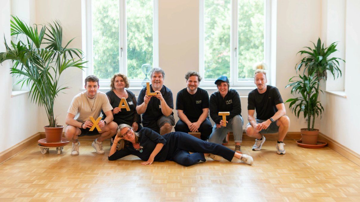 Sieben Teammitglieder in TEAM-XALT-Shirts halten in einem hellen Büroraum Holzbuchstaben in die Kamera