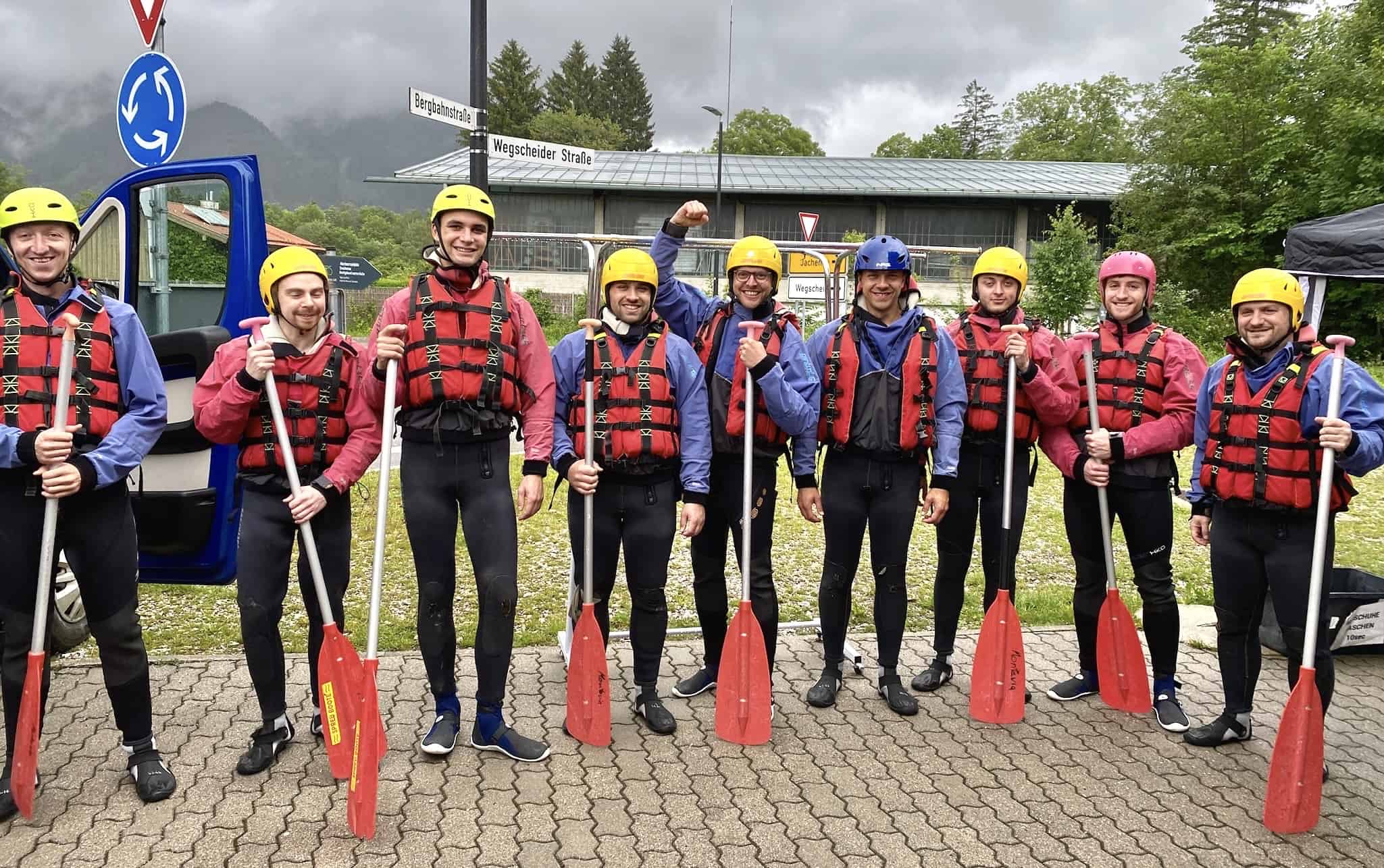 Neun Personen in Rettungswesten und Helmen mit Paddeln vor einem blauen Bus, Berglandschaft im Hintergrund.