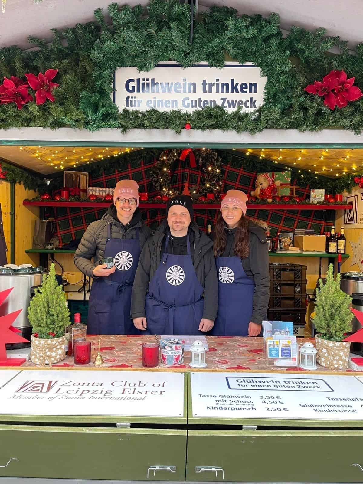Drei Personen in XALT-Schürzen und Mützen an einem Glühweinstand des Zonta Clubs Leipzig Elster mit Preistafel und Weihnachtsdekoration.