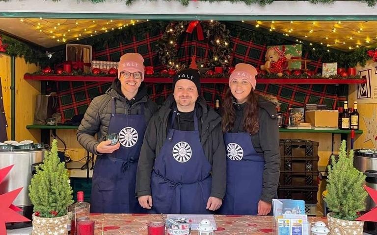 Drei Personen in XALT-Schürzen und Mützen an einem Glühweinstand des Zonta Clubs Leipzig Elster mit Preistafel und Weihnachtsdekoration.