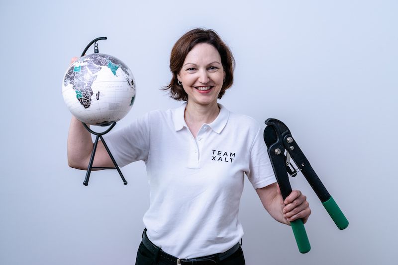 Eine lächelnde Frau in weißem Poloshirt hält einen kleinen Globus und eine Zange in die Kamera