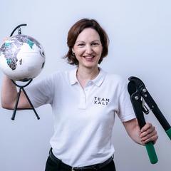 Eine lächelnde Frau in weißem Poloshirt hält einen kleinen Globus und eine Zange in die Kamera