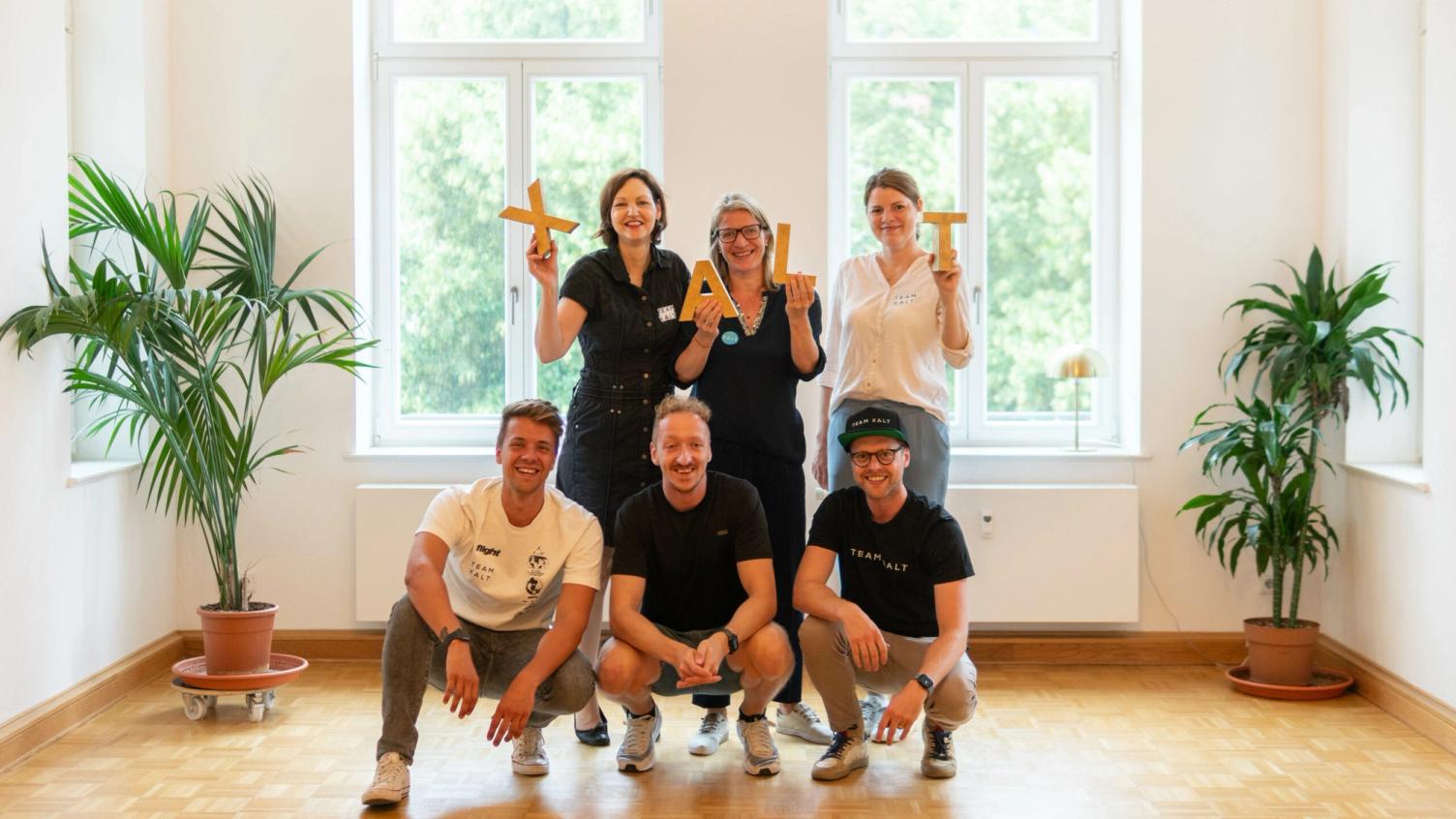 Sechs Teammitglieder posieren in einem hellen Raum mit großen Fenstern, drei hockend und drei stehend
