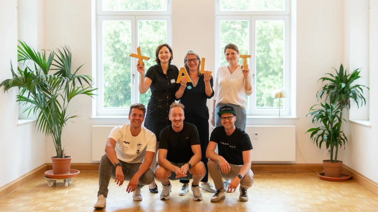Sechs Teammitglieder posieren in einem hellen Raum mit großen Fenstern, drei hockend und drei stehend