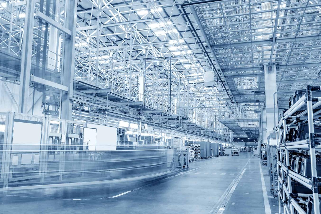 A long, tall manufacturing hall with steel scaffolding and overhead tracks; on the left a system passes by, blurred into streaks