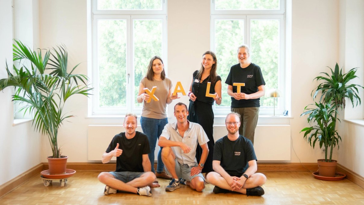 Sieben Teammitglieder posieren in einem hellen Büroraum mit Parkettboden und großen Fenstern