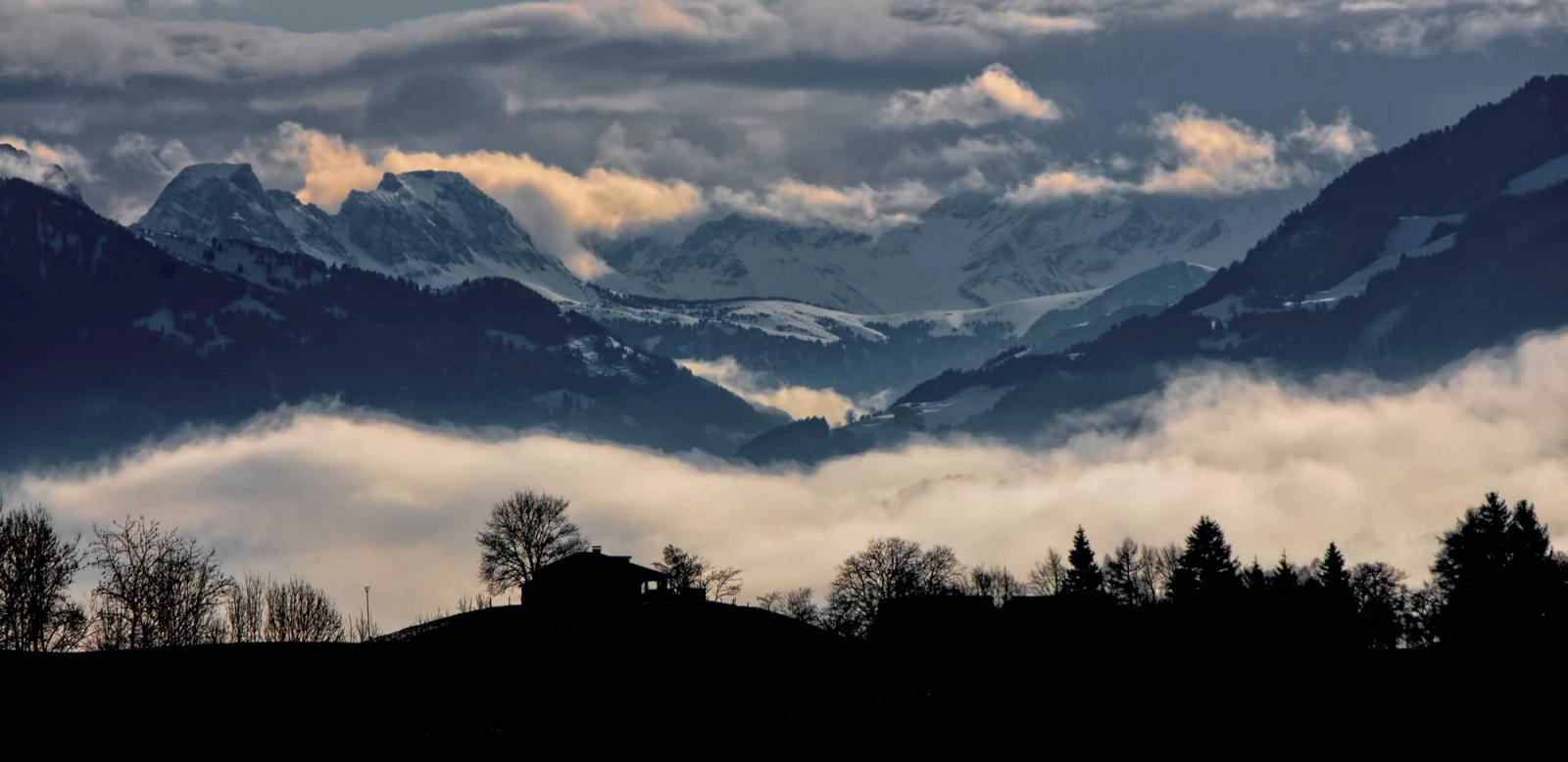 Alpenpanorama in der Dämmerung: schneebedeckte Gipfel über einem Wolkenmeer, davor die Silhouette einer Hütte auf einem Hügel