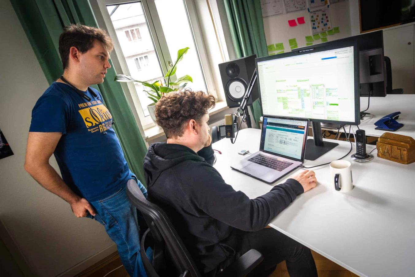 Two men work in an office at a desk with a laptop and monitor