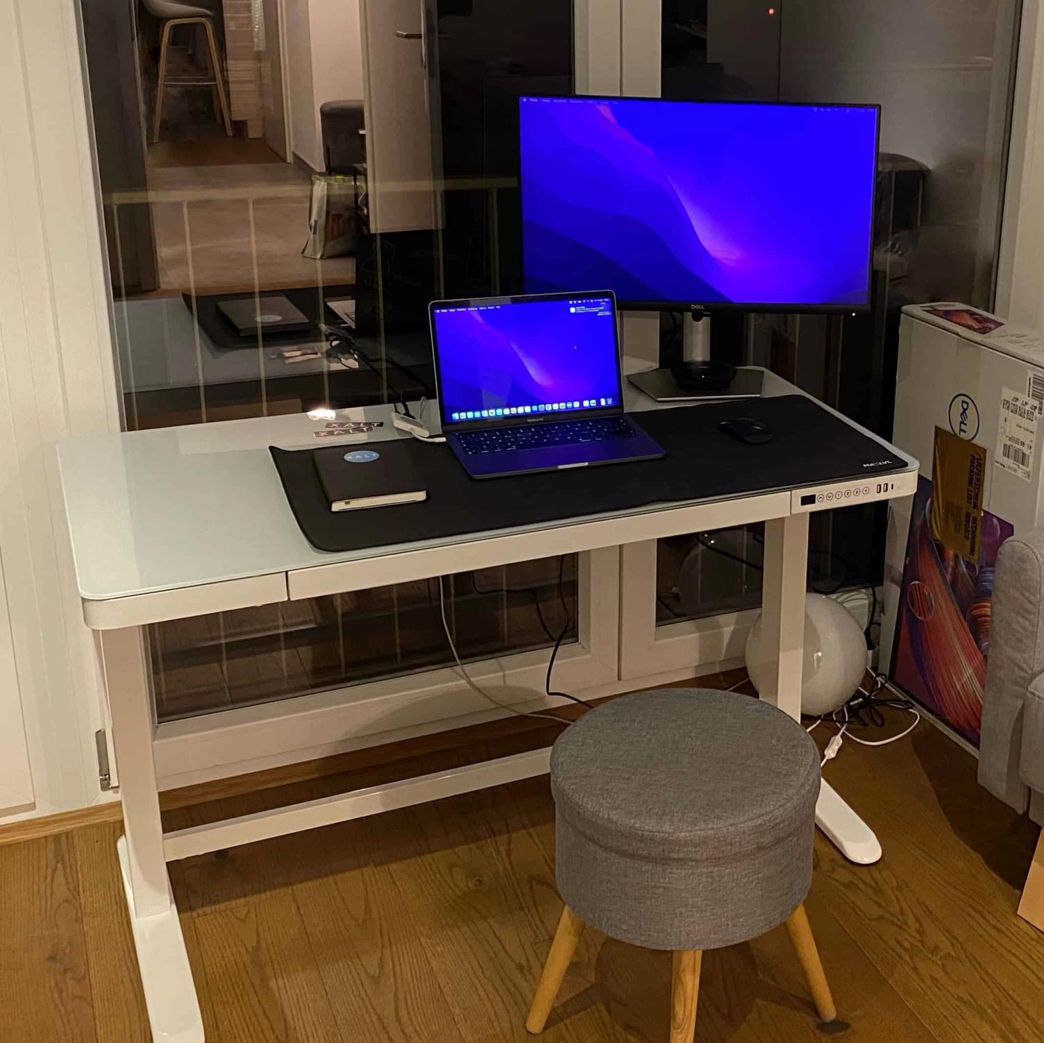 Foto eines höhenverstellbaren Schreibtischs am Fenster mit Laptop, zweitem Bildschirm, Schreibtischunterlage und einem gepolsterten Hocker
