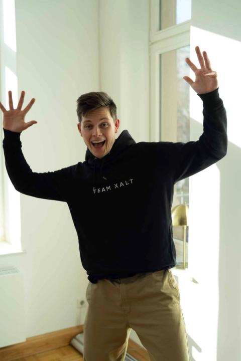 Ein Mann in schwarzem TEAM-XALT-Hoodie hebt in einem sonnigen Raum beide Arme