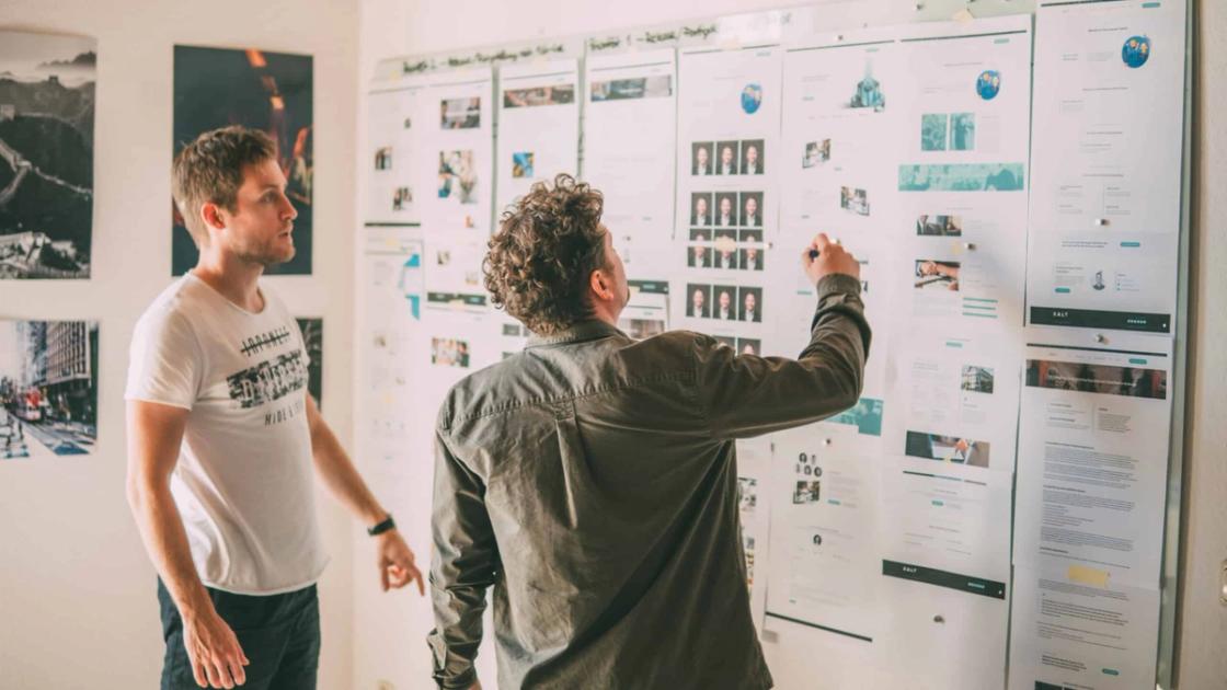 Zwei Männer stehen in einem Büro vor einer Wand mit ausgedruckten Web-Prototypen und Wireframes