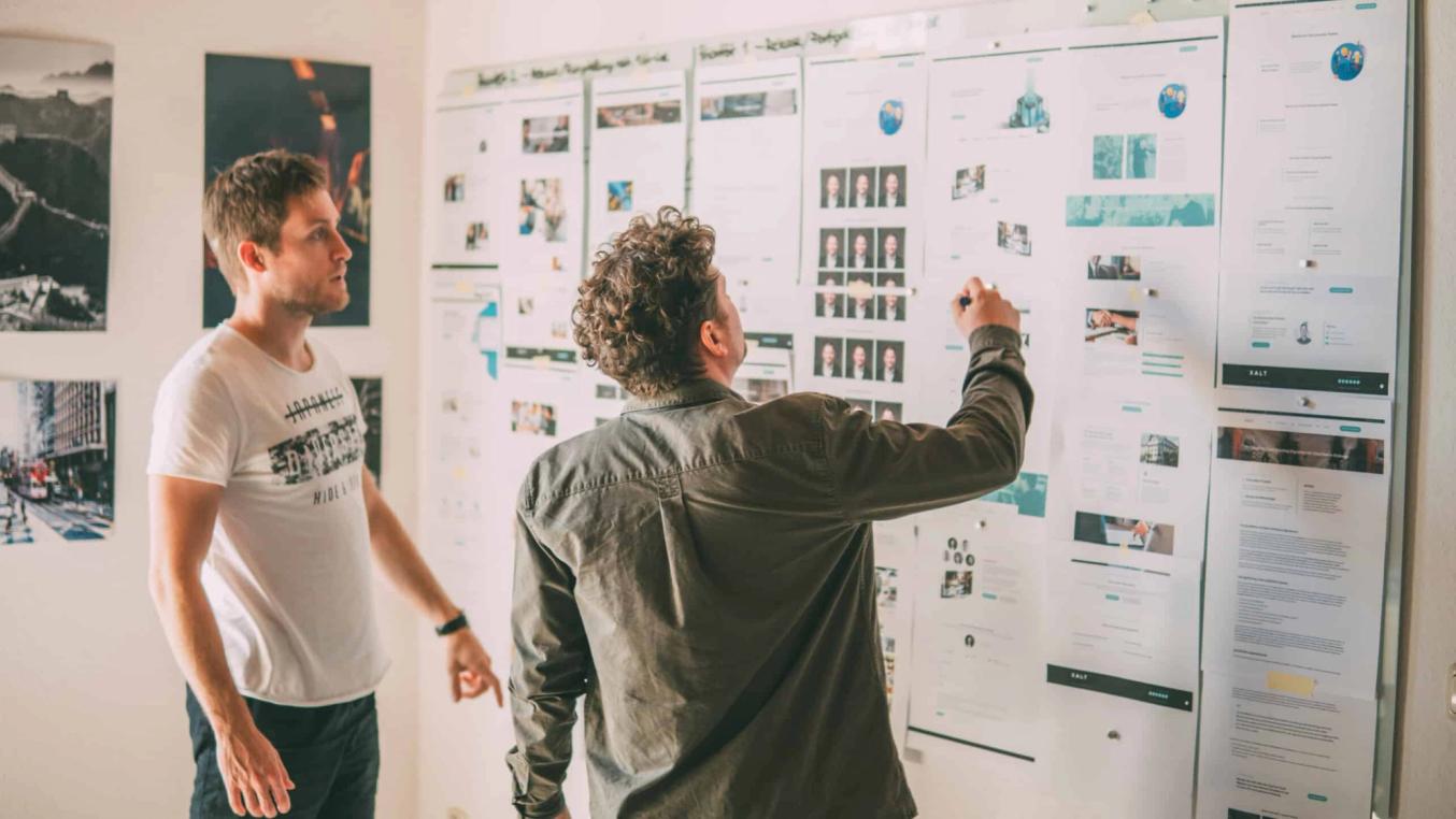 Zwei Männer stehen in einem Büro vor einer Wand mit ausgedruckten Web-Prototypen und Wireframes