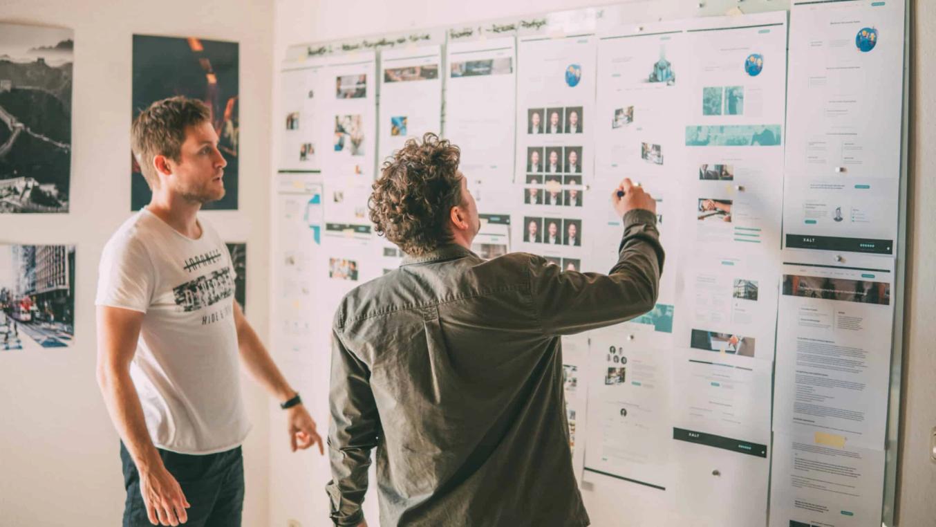 Zwei Männer stehen in einem Büro vor einer Wand mit ausgedruckten Web-Prototypen und Wireframes