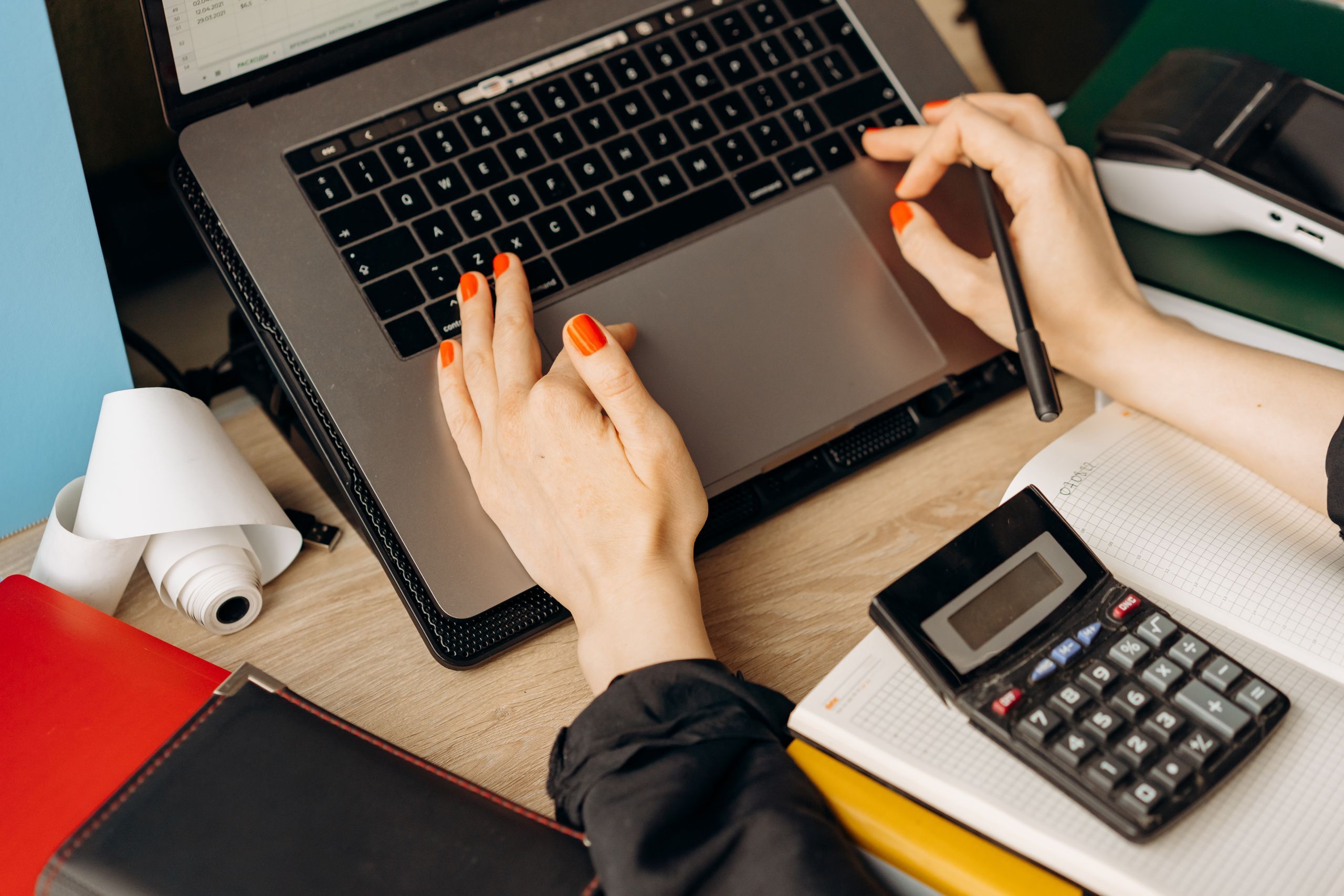 Hands on a laptop on a wooden desk, next to a calculator, a receipt roll and a notebook
