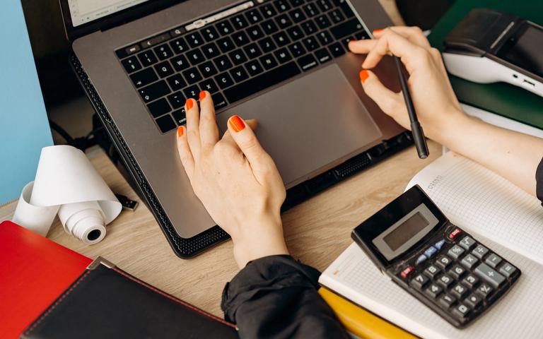 Hands on a laptop on a wooden desk, next to a calculator, a receipt roll and a notebook