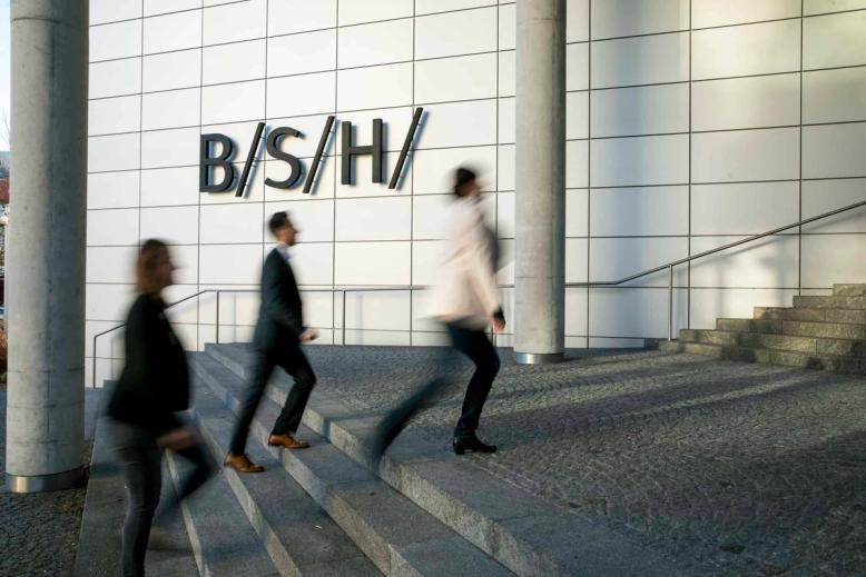 Eingang der BSH-Zentrale: drei Menschen gehen die Treppen hinauf, an der weissen Fassade das B/S/H/-Zeichen