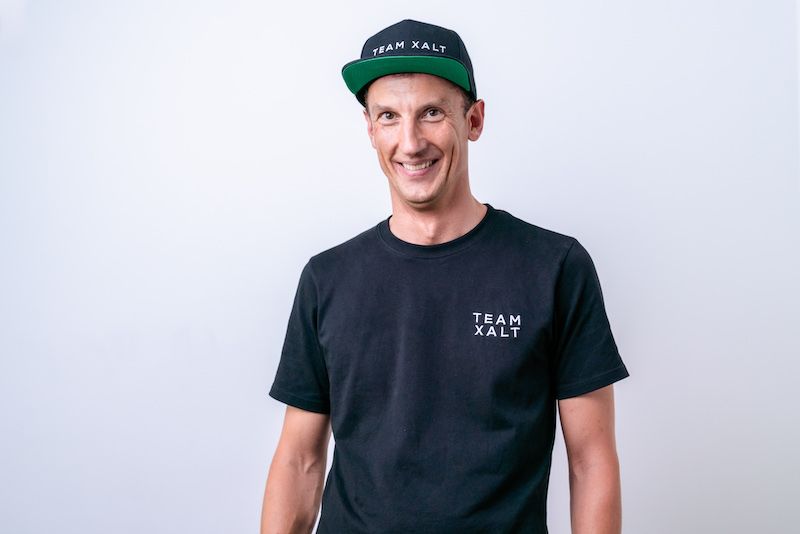 Ein lächelnder Mann mit schwarzer Kappe und TEAM-XALT-Shirt vor hellem Hintergrund