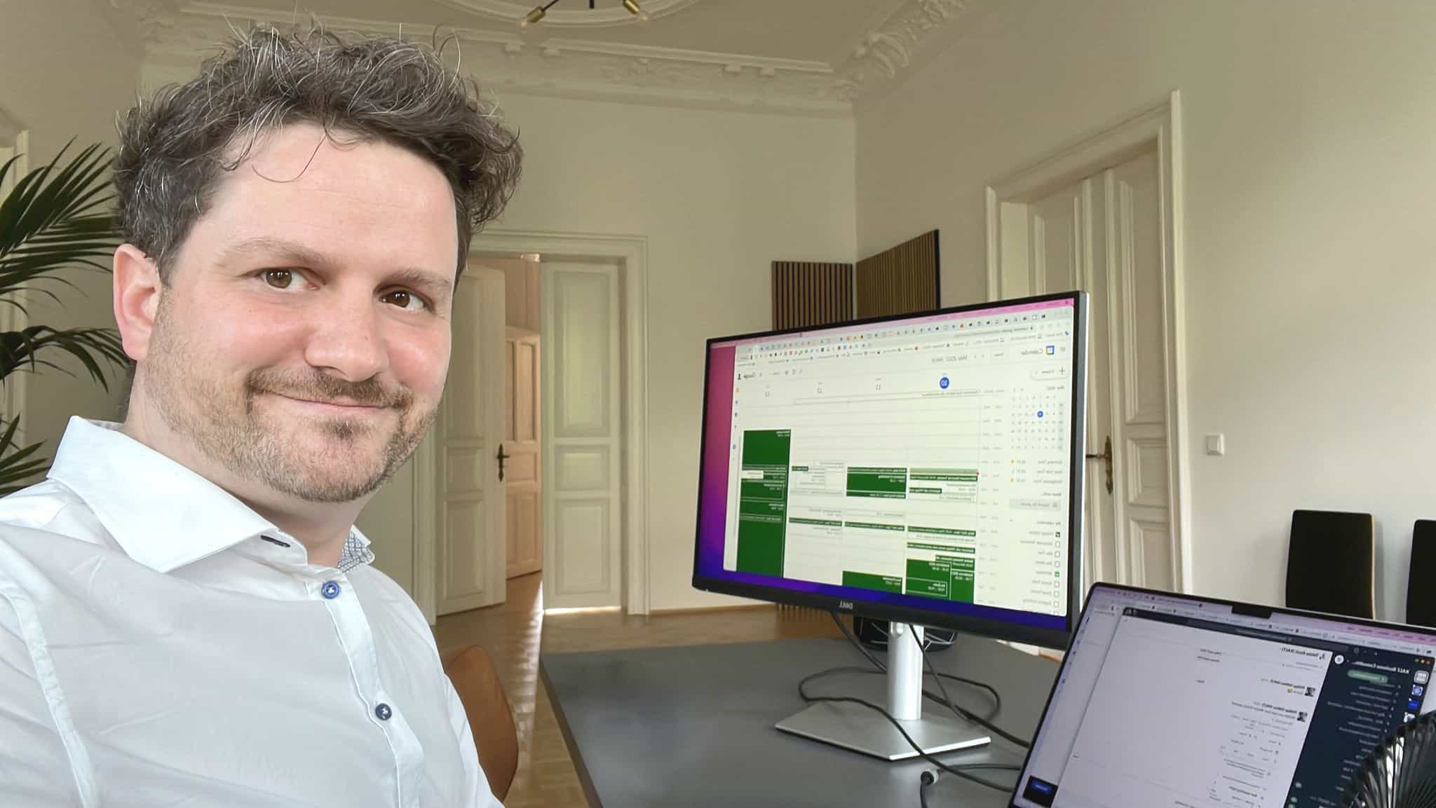 Mann im weißen Hemd vor Schreibtisch mit großem Monitor, auf dem ein Projektmanagement-Dashboard zu sehen ist.