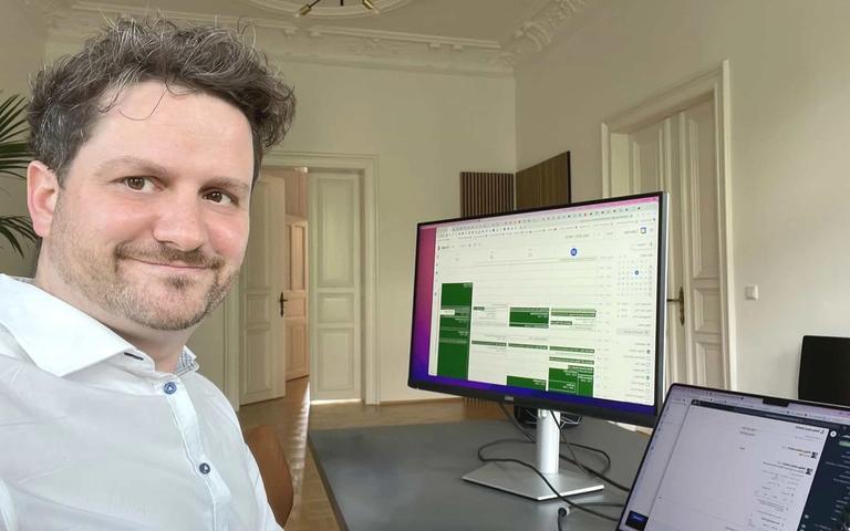 Mann im weißen Hemd vor Schreibtisch mit großem Monitor, auf dem ein Projektmanagement-Dashboard zu sehen ist.