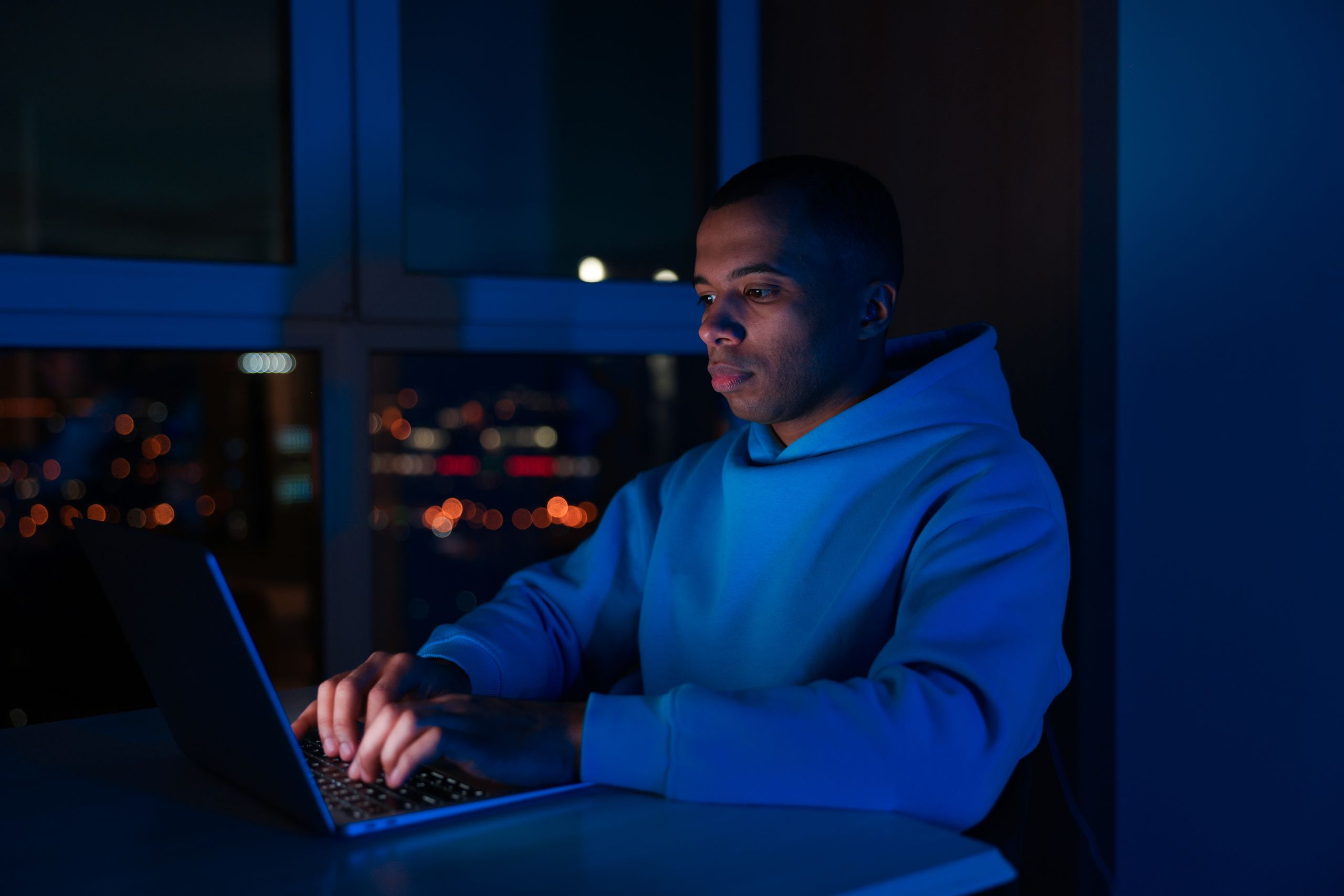 Man working on a laptop in a dark room at night, city lights in the background
