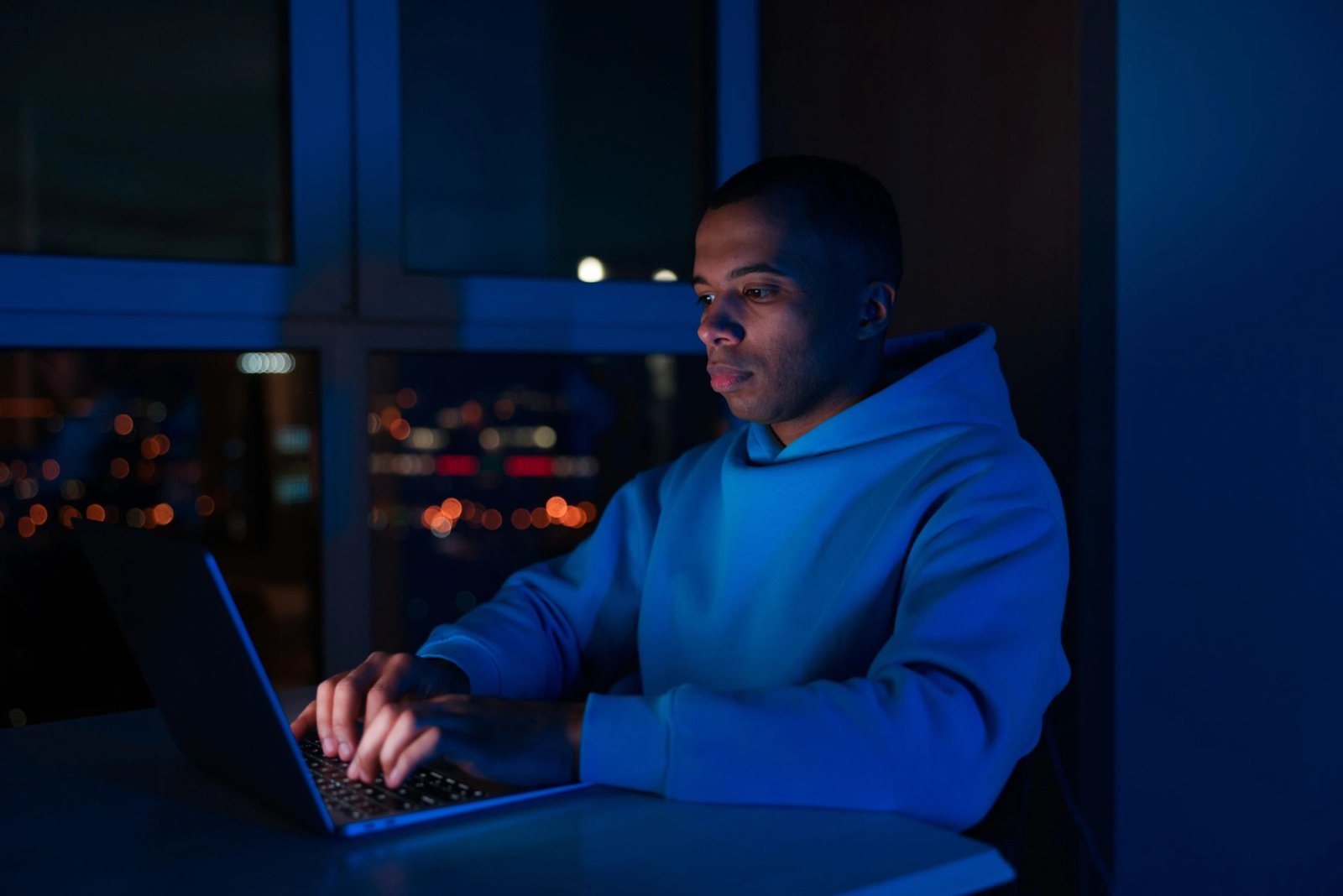 Mann arbeitet abends am Laptop in einem dunklen Raum, im Hintergrund die Lichter der Stadt