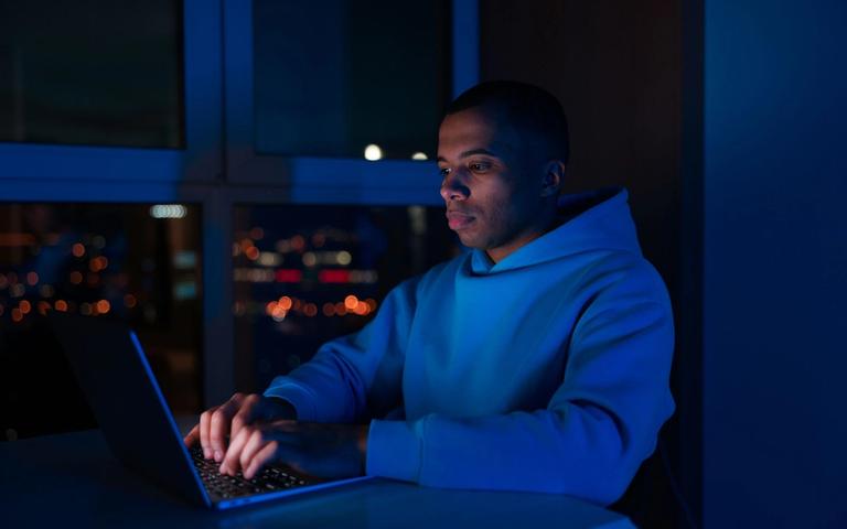 Man working on a laptop in a dark room at night, city lights in the background