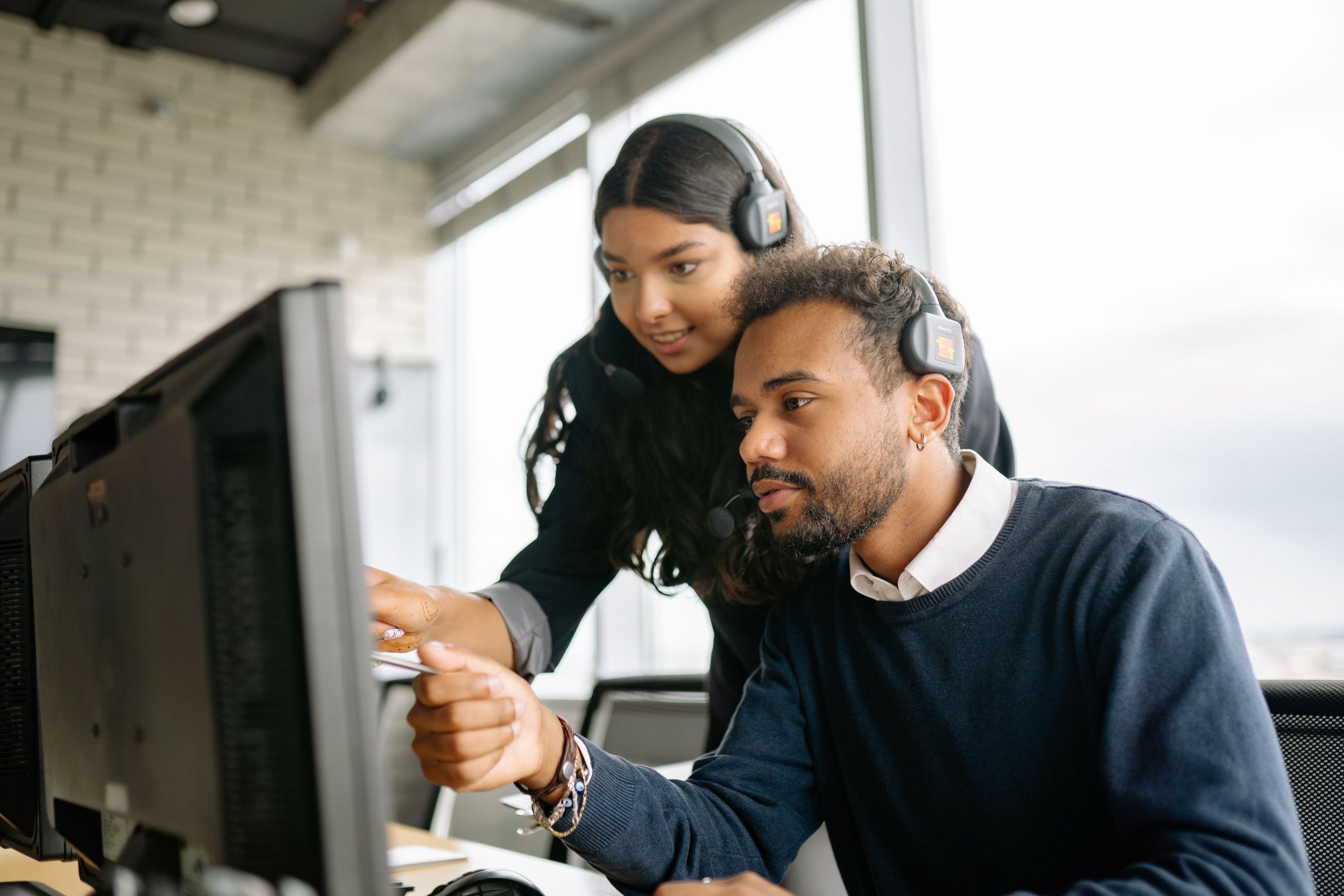 Two support agents wearing headsets discuss something at a monitor in a bright office