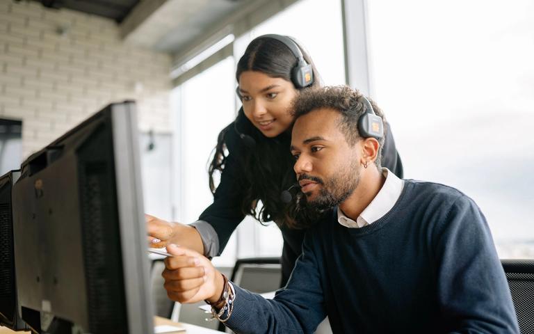 Two support agents wearing headsets discuss something at a monitor in a bright office