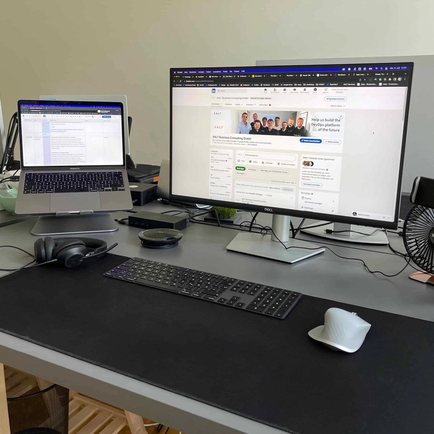 Foto eines Home-Office-Arbeitsplatzes: Laptop auf einem Ständer, großer Bildschirm mit der LinkedIn-Seite von XALT, Tastatur, Maus, Headset und Ventilator