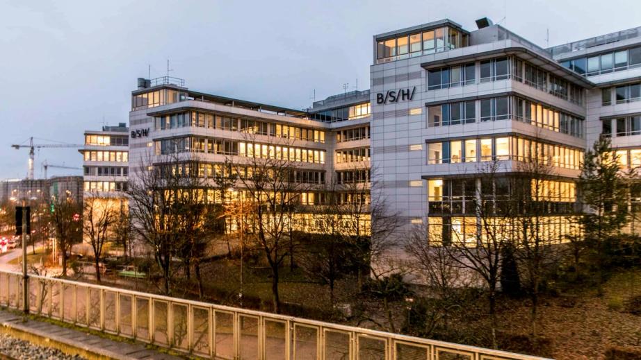 Die BSH-Zentrale in der Dämmerung: mehrere Bürogebäude mit erleuchteten Fenstern und dem B/S/H/-Zeichen an der Fassade