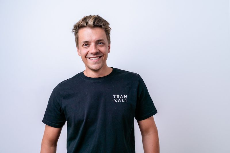 Ein lächelnder Mann mit blondem Haar in schwarzem TEAM-XALT-Shirt vor hellgrauem Hintergrund