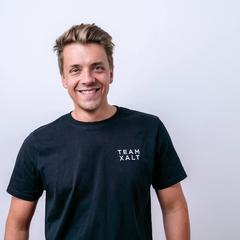 Ein lächelnder Mann mit blondem Haar in schwarzem TEAM-XALT-Shirt vor hellgrauem Hintergrund