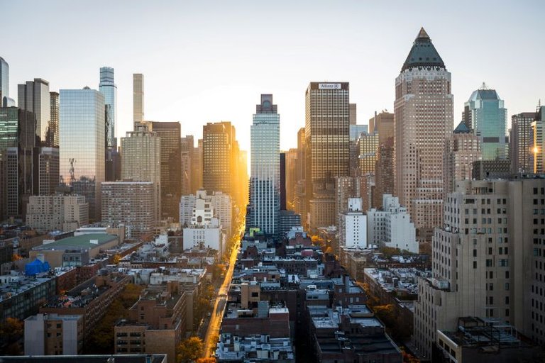 Eine Hochhauslandschaft von New York im Abendlicht, mit einer beleuchteten Straße zur Sonne hin