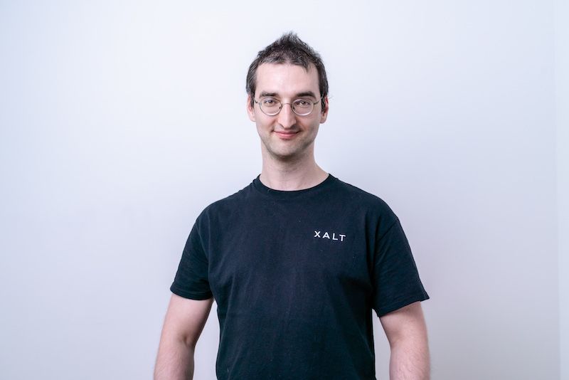 Ein Mann mit runder Brille in schwarzem XALT-Shirt vor weißem Hintergrund