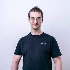 Ein Mann mit runder Brille in schwarzem XALT-Shirt vor weißem Hintergrund