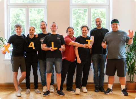 Acht Männer halten in einem hellen Raum gelbe Buchstaben, die das Wort XALT bilden