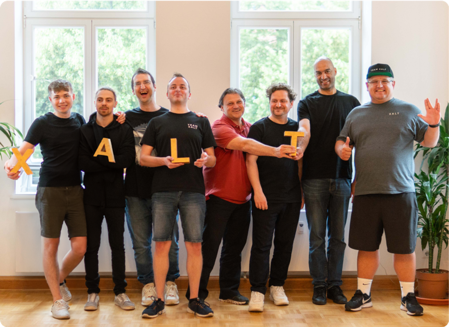 Eight men hold yellow letters spelling out XALT in a bright room