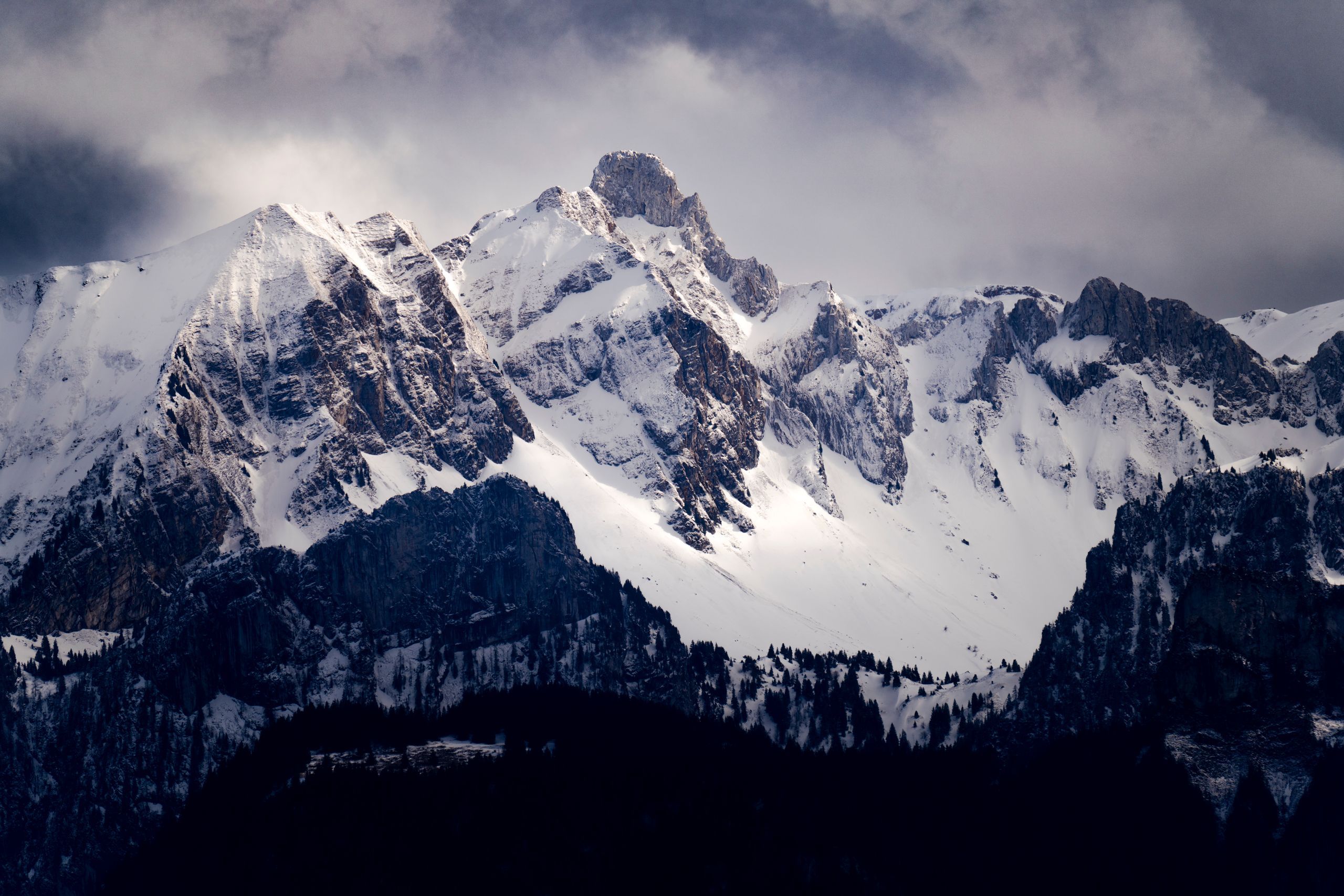 Snow-covered mountain range under a cloudy sky, with dark forested slopes in front