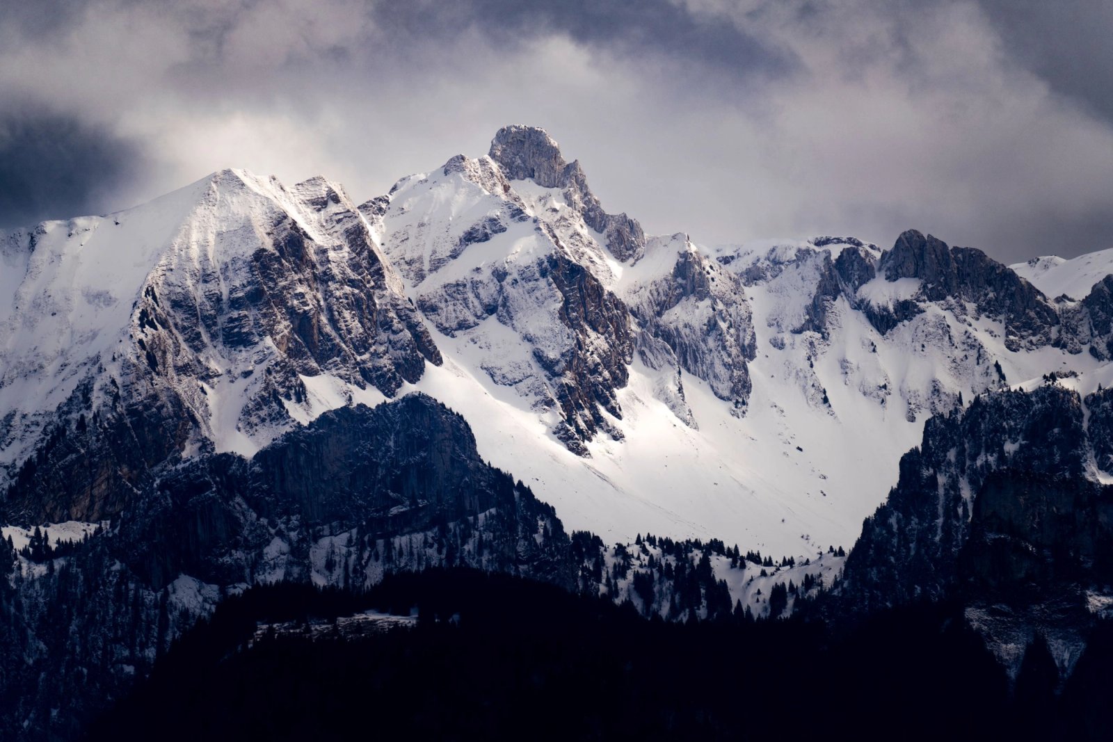 Snow-covered mountain range under a cloudy sky, with dark forested slopes in front
