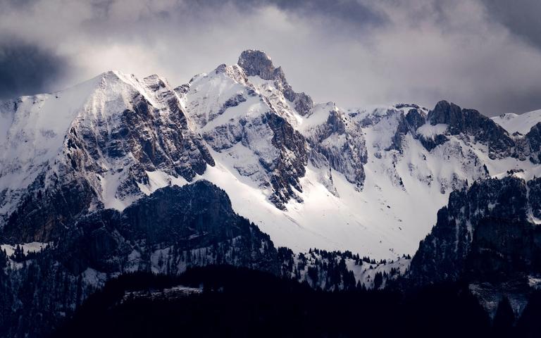 Snow-covered mountain range under a cloudy sky, with dark forested slopes in front