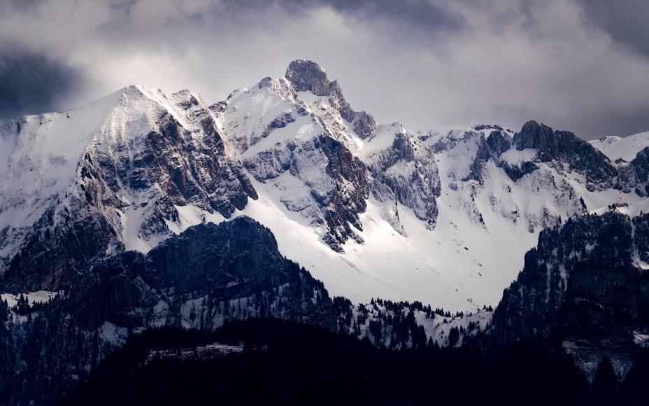 Schneebedeckte Gebirgskette unter wolkigem Himmel, davor dunkle bewaldete Hänge