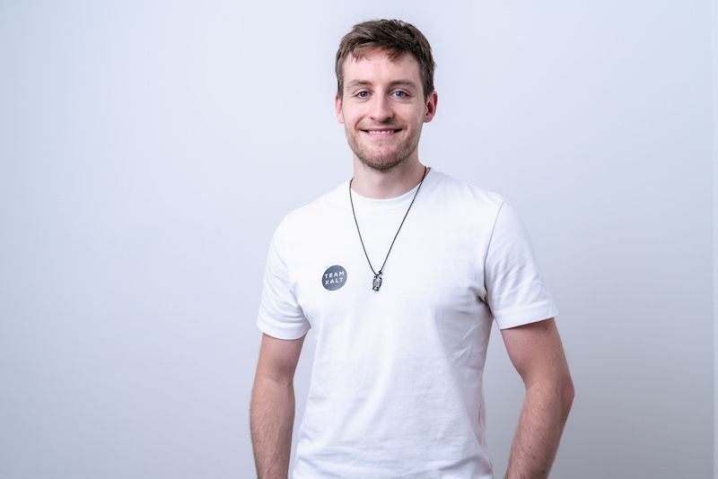 Ein lächelnder Mann in weißem Shirt mit rundem Aufnäher vor hellgrauem Hintergrund