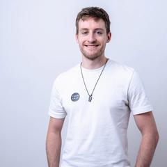Ein lächelnder Mann in weißem Shirt mit rundem Aufnäher vor hellgrauem Hintergrund